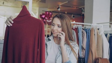 Cep telefonu üzerinde konuşurken ve modaya uygun giysiler Bayan giyim butik seçmek genç kadın. O örgü jumper arıyor, ona dokunmak ve gülümseyen.