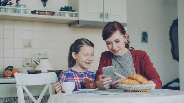 陽気な母と娘一緒にスマート フォンの閲覧と自宅の台所で朝の朝食時に笑みを浮かべて — ストック写真