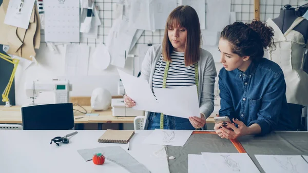 デザイン業界で働く魅力的な若い女性はスケッチを比較すると 現代のテーラー ショップで働いている間彼らの話 生産的なチームワークの概念 — ストック写真