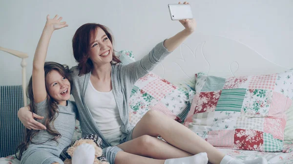 幸せな母と Selfie を撮っている女の子のスマート フォンのカメラで写真家で居心地の良いベッドルームに座りながらしかめっ面を楽しもう — ストック写真