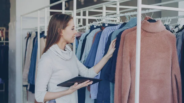 Young and successful owner of womens clothing shop is checking and counting garments on rails while using tablet. She is typing information about her goods. Small business concept.