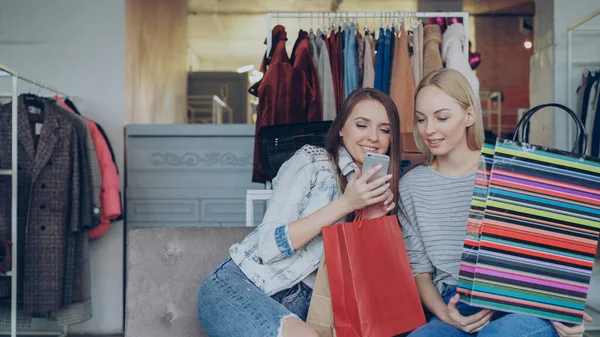 欢快的年轻妇女闲聊和使用智能手机 而坐在漂亮的服装精品店 他们微笑着 热情地示意 现代生活方式概念 — 图库照片