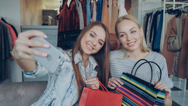 Close-up shot of two young women making selfie with smart phone after shopping in womens clothing store. First they are posing with paper bags and laughing, then watching photos on screen