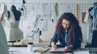 Genç kadın moda tasarımcısı modern atölye masasında bayanlar elbise kroki çizim. Manken, dikiş öğeleri, gitmek kahve görünür. Giyim üretim kavramı yaratıcılık.
