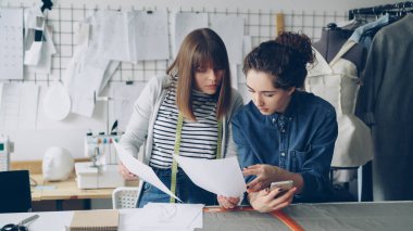 Yaratıcı kadın moda tasarımcıları dikiş Resepsiyon çalışma çizimleri izlerken meşgul. Genç kadınlar konuşmaya ve smartphone kontrol. Modern terziler Dükkanı arka planda.