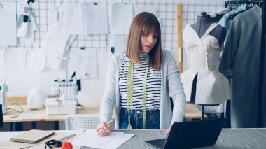 Güzel moda tasarımcısı giyim çizimler onun yeni elbise koleksiyonu için çizim ve laptop ile çalışma. Işık stüdyo ile Rails giysiler ve arka planda duvar çizimleri.