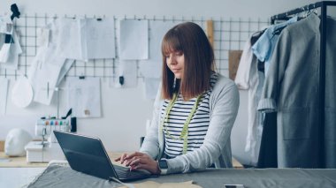 Genç serbest meslek terzi dizüstü kullanarak onu müşterilerle iletişim kurma. Arka planda ışık modern stüdyo iyi görünüm. Giyim tasarım sanayi kavram başarılı e-ticaret.