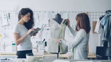 Onun iş arkadaşı ölçüm verileri yazmak için tablet ile çalışırken profesyonel terzi ölçü-bant ile manken üzerinde desenler ölçmek. Arka planda hafif modern atölye.