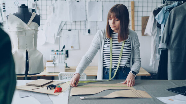 Young pretty seamstress is measuring clothing patterns with tape-measure at studio table while working in her modern tailors shop. Getting ready for sewing clothes concept.