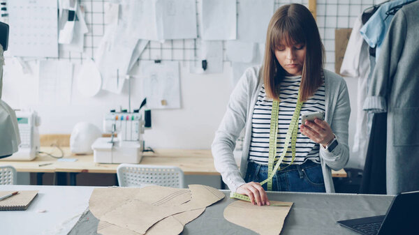 Clothing design entrepreneur is checking paper sewing drafts and looking at smartphone. She is examining every cutout and thinking about future garment. Modern technology in sewing industry concept.