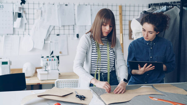 Female clothing designer is drawing outlines of new garment on fabric with chalk while her colleague is helping her and using tablet. Modern technologies in clothes manufacturing concept.