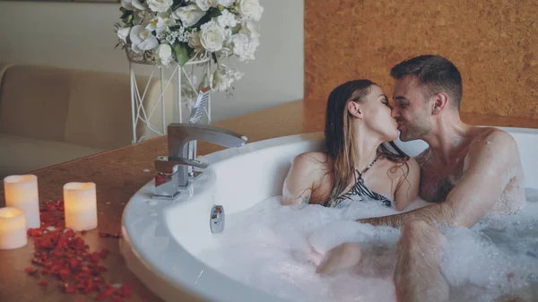 迷人的快乐情侣在热浴盆里玩吹泡沫 亲吻和享受浪漫的时刻 玫瑰花瓣 美丽的花朵和燃烧的蜡烛是可见的 — 图库照片