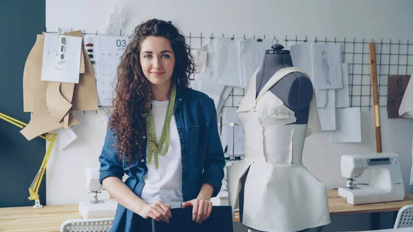 Waist-up portrait of beautiful young woman tailor standing in workplace near clothed mannequin and looking at camera. Womens garments, sketches and sewing machines are visible.