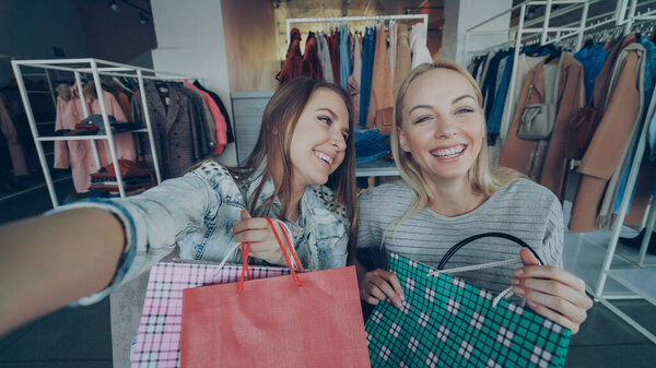 Point of view shot of two beautiful young women making selfie together with coloured paper bags in womens clothing department. Girls are posing, laughing and chatting happily