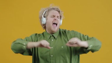 Kulaklıkla müzik dinleyen, sarı arka planda şarkı söyleyen ve dans eden yaratıcı adamın yavaş çekim portresi. İnsanlar ve eğlence konsepti.