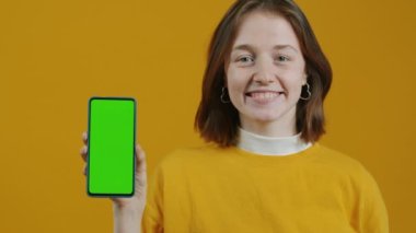 Krom anahtar yeşil ekranlı akıllı telefonu işaret eden ve sarı arka planda gülümseyen mutlu öğrencinin yavaş çekimde portresi. Aygıtlar ve çokluortam kavramı.