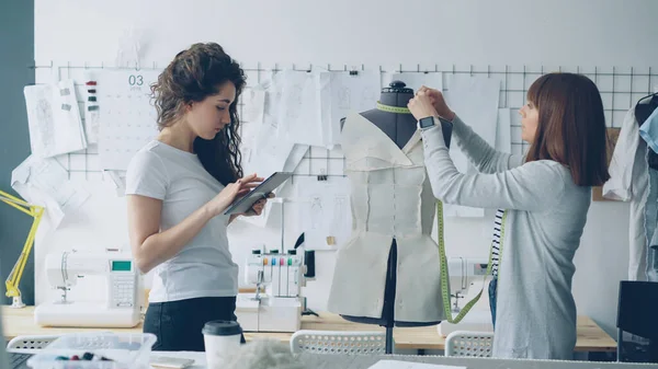 Clothing designer entrepreneurs are measuring mannequin parts while working together in nice modern studio. Young attractive woman is using tablet to enter measurement results.