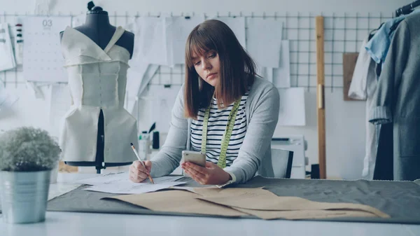 創造的な服のデザイナーはスマート フォンの画面を見て ミシン テーブルにモダンなテーラー ショップで働きながらスケッチを描画します 女性は忙しいとプロセスに関与します — ストック写真