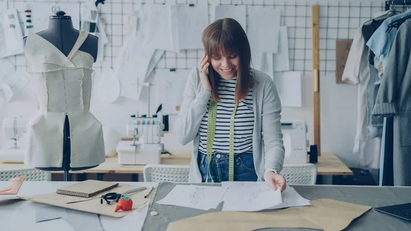 Young attractive dressmaker is discussing clothing designs on mobile phone and checking sketches while working in her modern studio between dummy and rail with clothes.