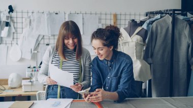 Yaratıcı terziler smartphone kullanarak ve giyim skeçler güzel modern stüdyoda çalışırken arıyorum. Kukla, dikiş makinesi ve kadın giysiler içinde arka plan.