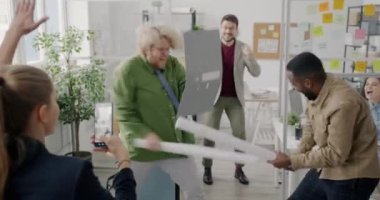 Çalışan oyuncular kağıt kılıçlarla savaşırken, kadın meslektaşları akıllı telefonlarıyla video kaydediyor.