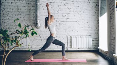 Genç ince sarışın kız beyaz duvarlı güzel Studio karmaşık yoga yapıyor. O savaşçı pozlar ile başlayan daha sonra ileri ve geri bükme ve namaste ile bitirme.