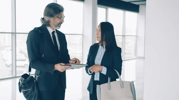 Elegant Asian woman is signing business contract and talking to partner mature Caucasian man indoors in commercial building. Real estate and deals concept.