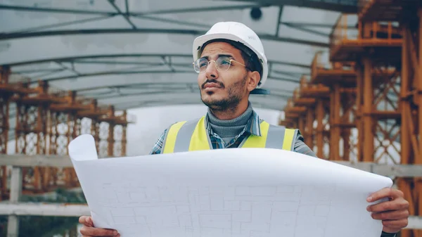 Portrait of Middle Eastern construction employee wearing uniform studying blueprint outdoors in building area. Commercial projects and workers concept.