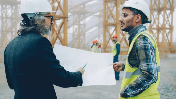 測量士が設備を背景に忙しい中 設計図を見て建設プロジェクトを議論するエンジニアのバックビュー 建築及び経営の考え方 — ストック写真