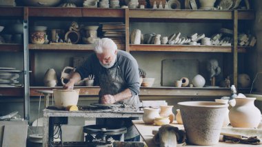 Çalışkan gümüş saçlı adam kil ile işyerinde, potters tekerlek üzerinde kerpiç parça şekillendirme çalışıyor. Güzel seramik mutfak eşyaları, el yapımı tencere ve raflarda vazolar görülebilir.