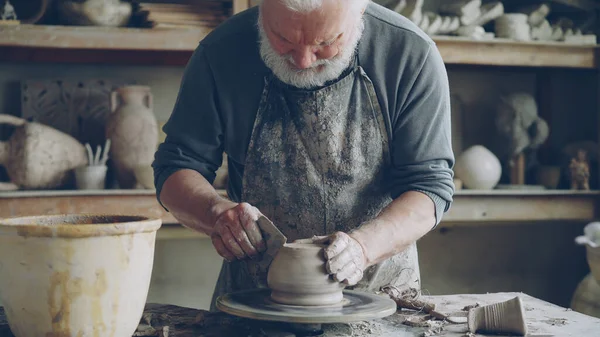 Senior male potter is working at bottom part of ceramic pot on spinning potters wheel in workplace. Creating traditional eathenware and conventional pottery concept.