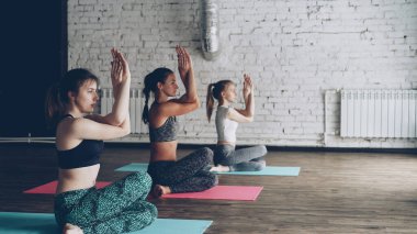 Kadın yoga eğitmeni inek yüz poz Gomukhasana onu dinlerken ve bu pozisyonda çalışan öğrenciler ise silah büküm sergiliyor. Genç kadın yan görünüm beyaz duvarlı stüdyom.
