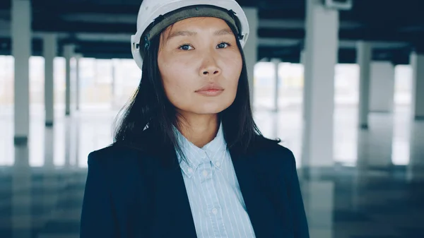 Portrait of elegant Asian business lady wearing protective helmet standing inside empty plant buidling in daytime ready for rent or sale deal