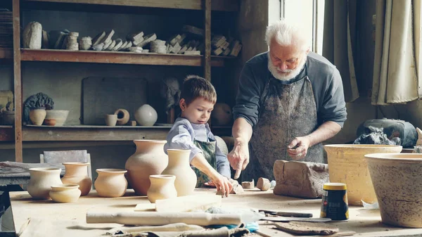 Cheerful little boy is throwing pieces of clay on work table while helping his loving grandfather in potters workshop. Happy childhood, family traditions and hobby concept.