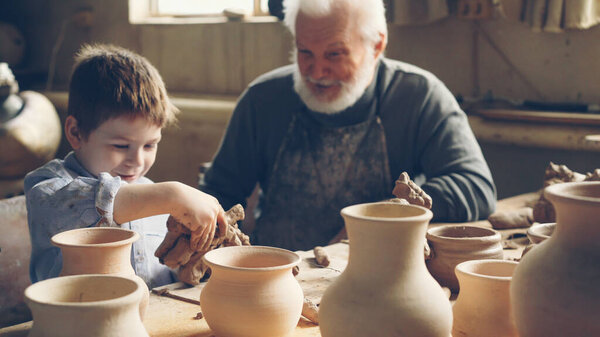 Cute little boy is playing with clay sitting with his grandparent at table in home pottery studio. Grandfather is talking to curious child, boy is enjoying new hobby.