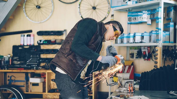 Experienced maintenance man is using electric circular saw while working in his small workplace. Young man is wearing protective glasses and gloves and is listening to music with earphones.