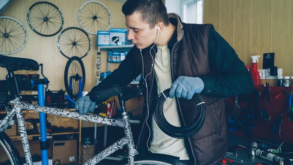 Young mechanic owner of bike repairing workshop is fixing broken bicycle holding bundle of wire and fixing it to bike frame. Small business and maintenance concept.