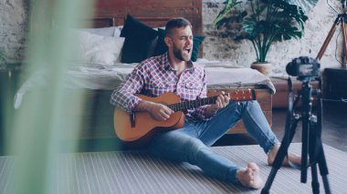 Genç yakışıklı müzisyen popüler blogger video iskambil gitar ve şarkı şarkı yatak odasında katta oturan kamera ile kayıt olan. Bloglama ve müzik konsepti.