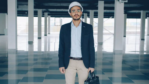 Portrait of ambitious construction investor in helmet and suit standing in empty plant building and looking at camera. Commercial property and investment concept.