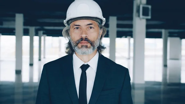 Portrait of experienced male architect standing inside new modern building wearing safety helmet and suit. Commercial property and architecture concept.