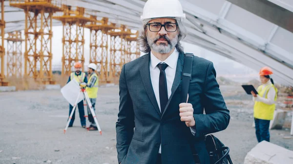 portrait of construction project manager standing outdoors wearing helmet and formal suit while workers are busy in background