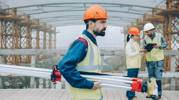 バックグラウンドで作業している間に機器を運ぶ建設現場外を歩く安全制服を着たプロの測量士の肖像 — ストック写真