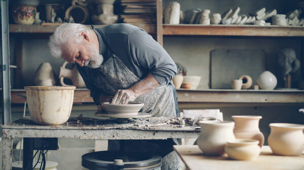 Experienced senior master is making low clay bowl on a pottery wheel in small home workshop. Ceramics jigger, handmade clayware and potters tools are visible.