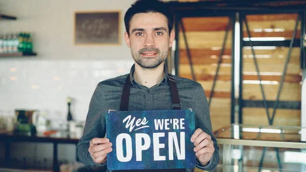 陽気なハンサムな男性の小さなコーヒーハウスの所有者は 近代的なコーヒーショップの中に立っている間 オープンサインを保持している 新しいビジネスや人々の概念を開く — ストック写真