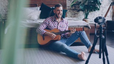 Genç yakışıklı müzisyen popüler blogger video iskambil gitar ve şarkı şarkı yatak odasında katta oturan kamera ile kayıt olan. Bloglama ve müzik konsepti.