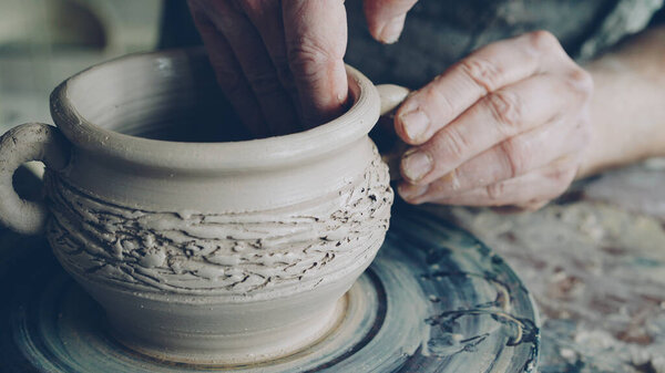 Close-up shot of muddy male potters hands shaping and fixing ears of ceramic bowl on throwing wheel. Nimble craftsman is forming clay professionally creating beautiful clayware.