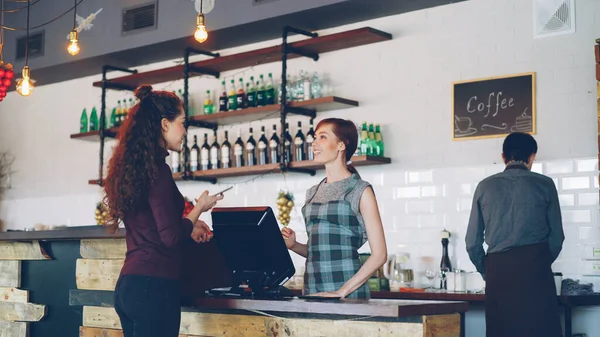 Young people are buying coffee-to-go in nice local cafe and paying with smartphone while friendly workers are greeting customers, talking and selling drinks.