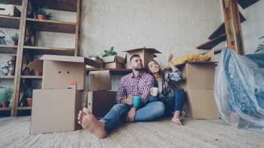 Sevimli genç çift, taşındıktan sonra yeni dairenin zemininde otururken konuşuyor ve kupa tutuyor. Sayısız kutu, paketlenmiş mobilya ve bitkiler görünür..