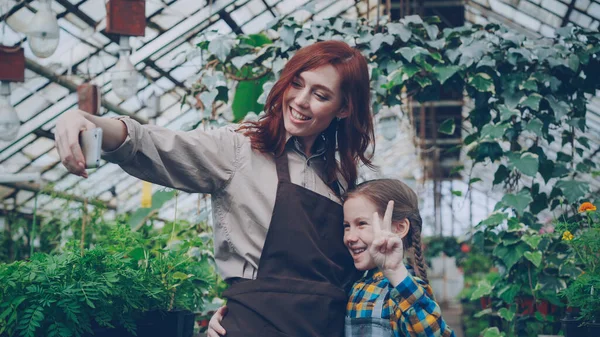 温室効果のきれいな女性オーナーとかわいい娘はポーズと笑顔 果樹園の中のスマート フォンで Selfie を取っています 人と現代の技術の概念 — ストック写真