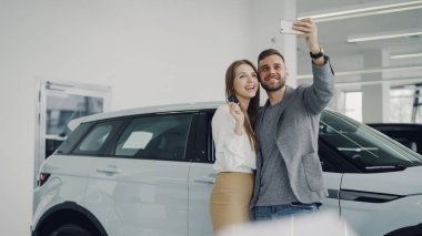 Çekici genç çift selfie arabanın anahtarlarını modern motor showroom ile yeni otomobil satın alma sonra alıyor. Bunlar kameraya bakarak, poz, Öpüşme ve gülümseyen.